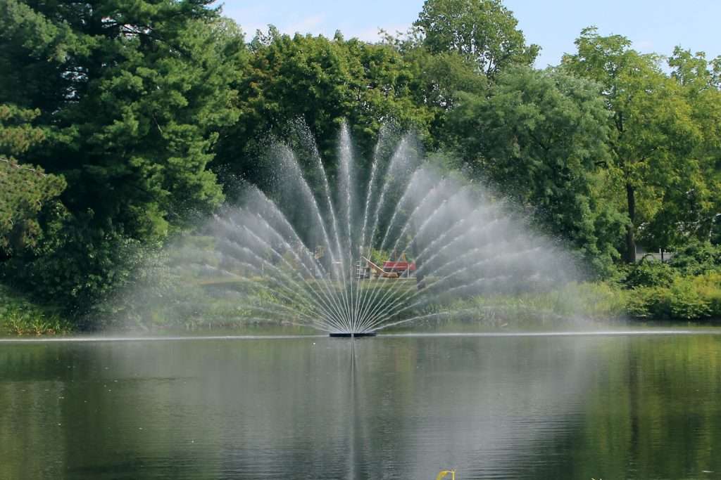 Otterbine Aries fountain in the middle of a lake with tall spray patterns and trees in the background