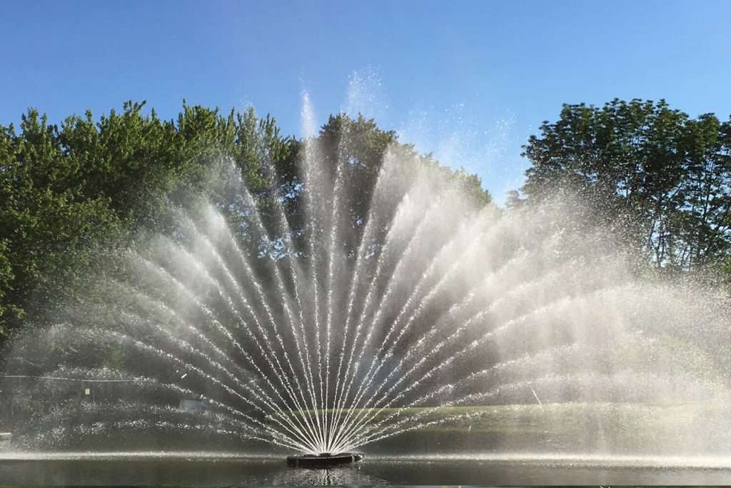 Otterbine Aries fountain producing a wide fan-like spray of water in a pond under blue sky