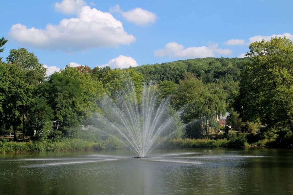 Otterbine Equinox fountain spraying water in a lake surrounded by lush green trees and hills under a blue sky