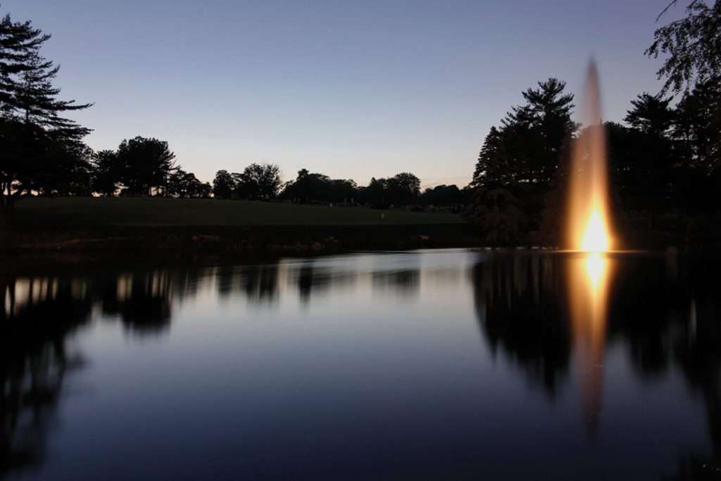 Otterbine Mystic fountain illuminated at night, glowing over a still lake with trees silhouetted in the background
