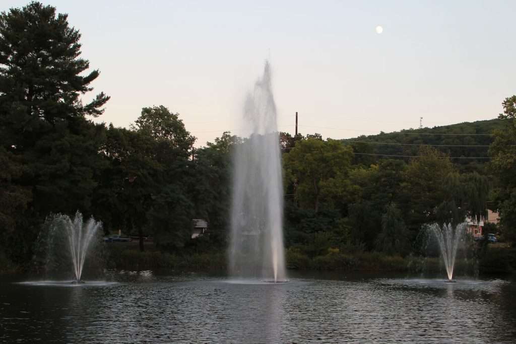 Otterbine Mystic fountains spraying water in a lake at dusk with moon visible above trees