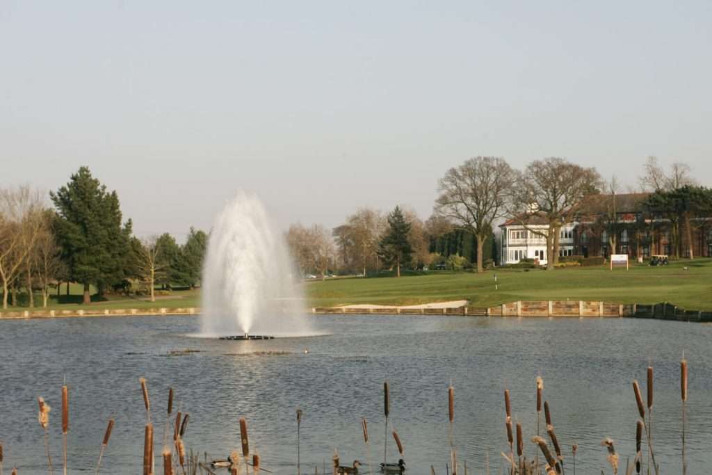 Otterbine Polaris fountain spraying in a park lake with reeds in the foreground and clubhouse in the distance