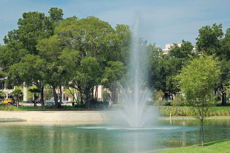 Otterbine Triad fountain in a clear pond bordered by trees and a park setting