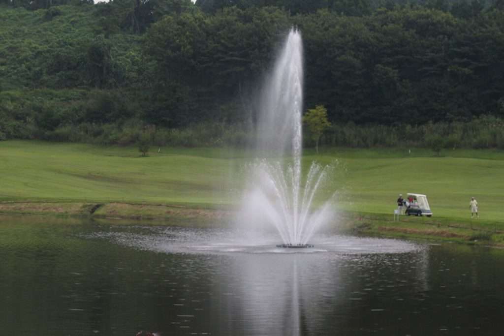 Otterbine Triad fountain in a pond with golfers and a golf cart in the background