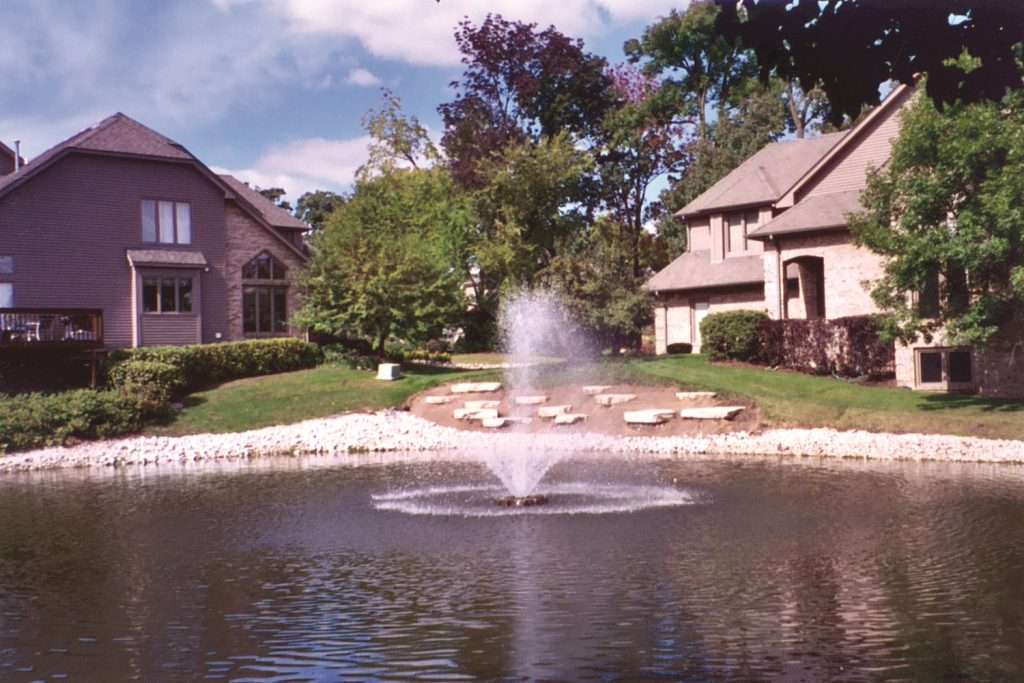 Fountain in pond with suburban houses and trees in background.
