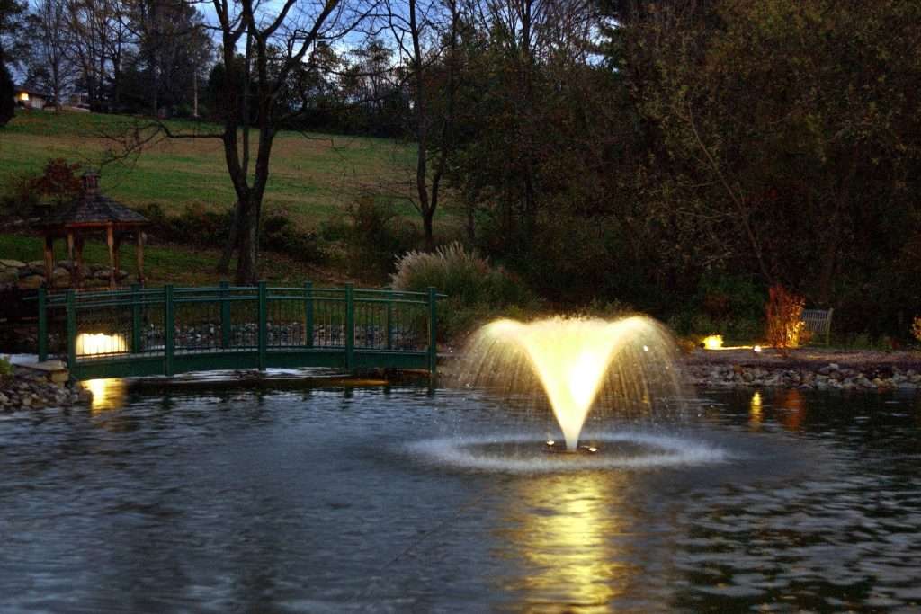 Illuminated fountain in pond near green bridge at dusk.