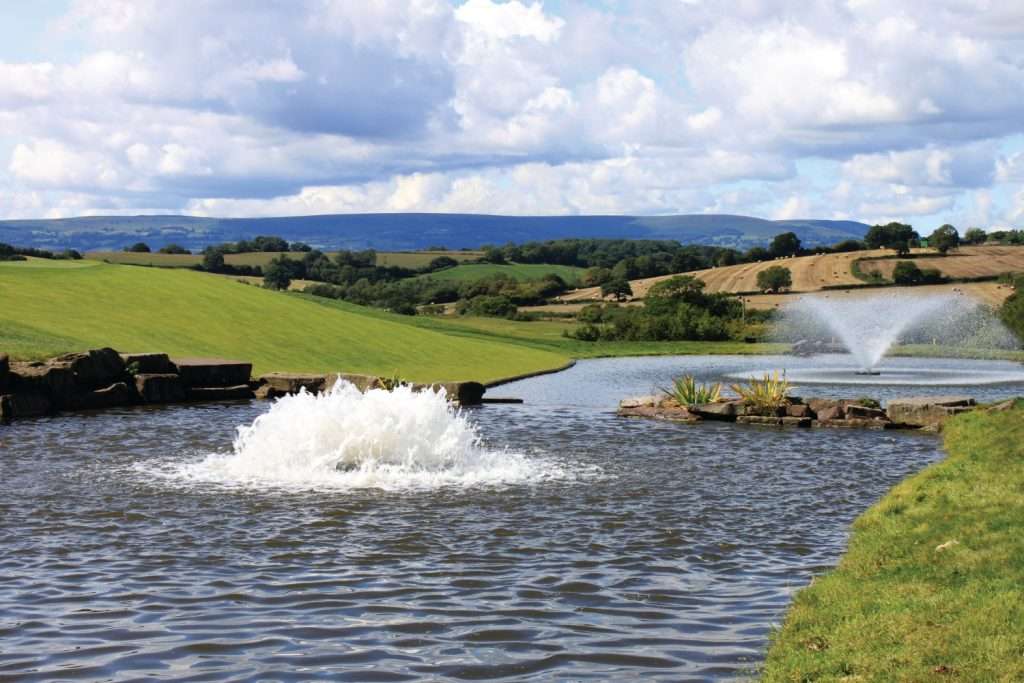 Scenic pond with fountains and rolling green hills under cloudy sky.
