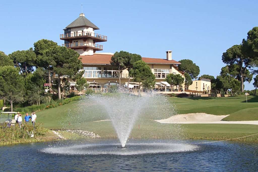 Golf course with clubhouse, fountain, and trees in the background.