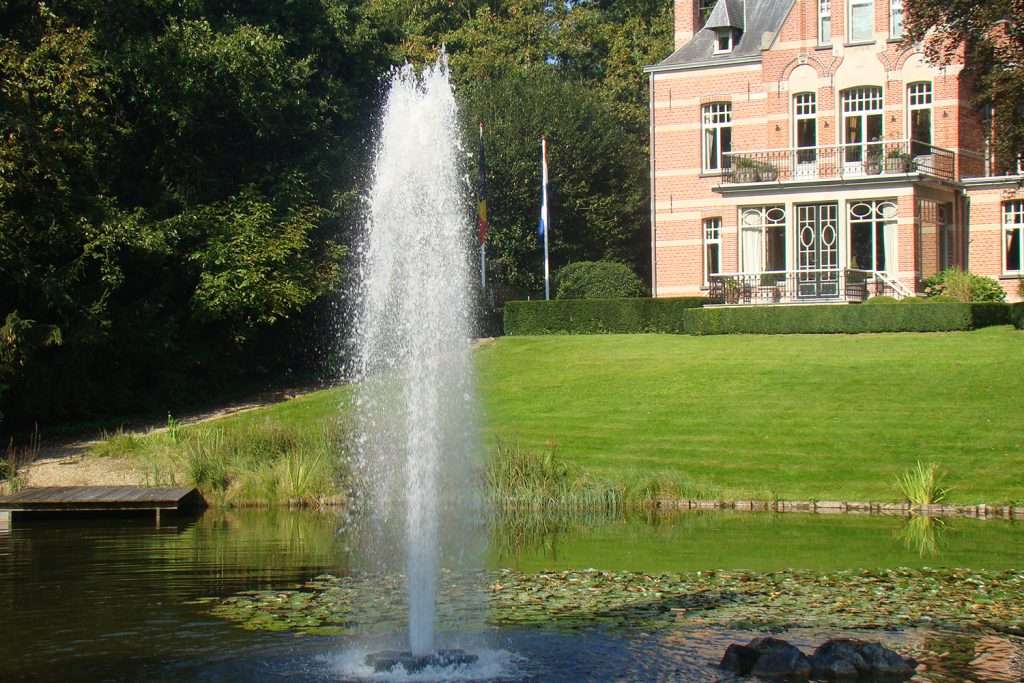 Fountain in front of brick mansion with lush green lawn.