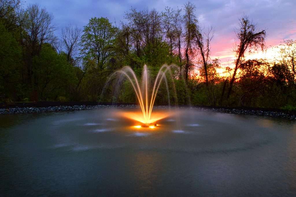 Illuminated fountain with vibrant sunset and trees in the background.