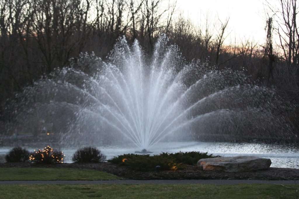 Large water fountain spraying in pond, surrounded by trees.