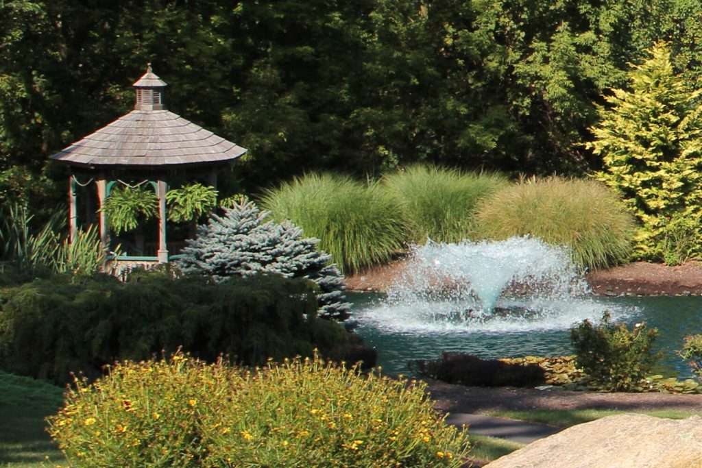 Garden gazebo beside pond with fountain and lush greenery.
