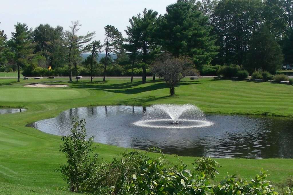 Golf course with pond and fountain, surrounded by trees.