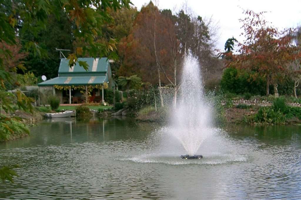 Otterbine Tristar fountain in a pond with a cottage and autumn trees in the background