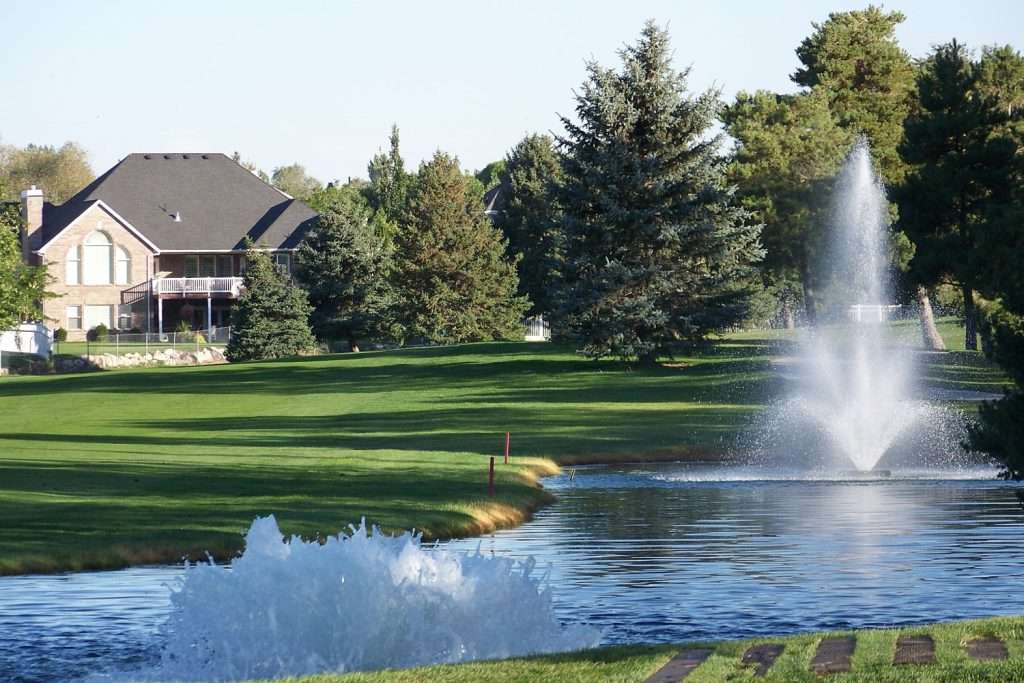 Golf course with pond and fountains, surrounded by trees and houses.