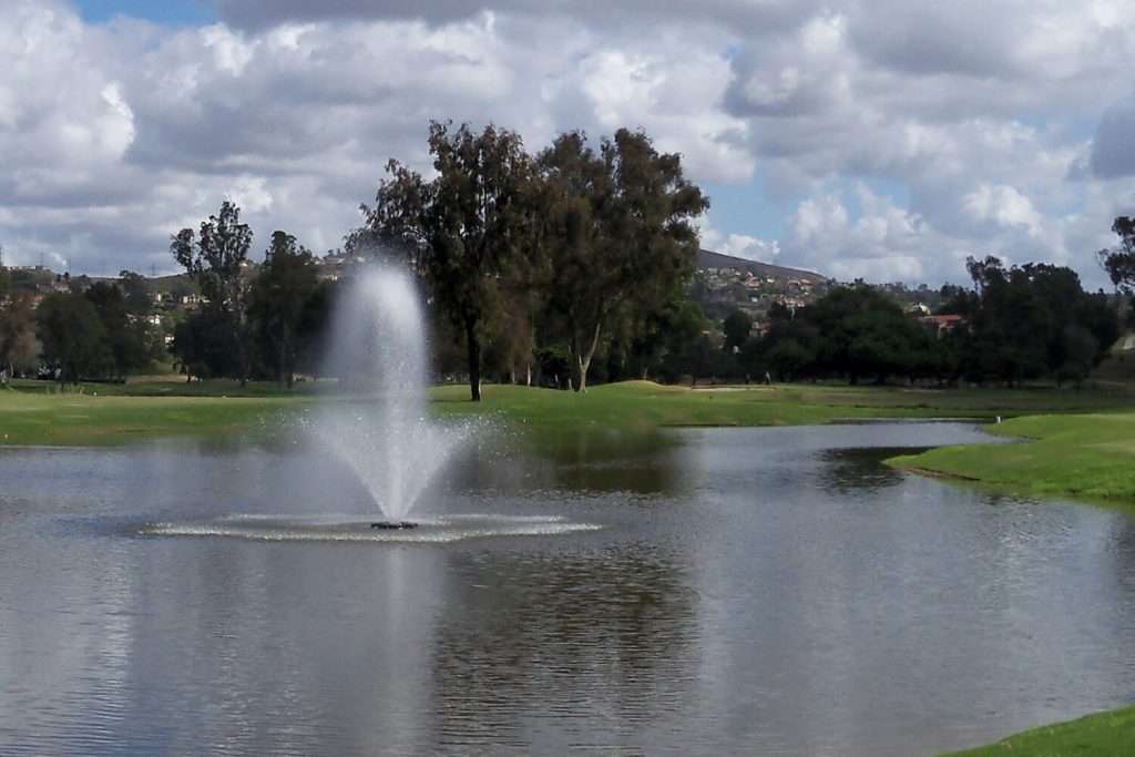 Fountain in pond with trees and cloudy sky.